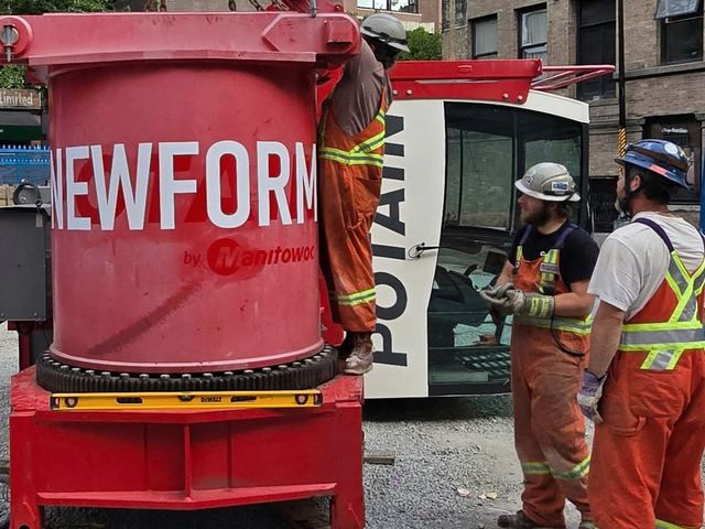 Three Newform crew members in high-visibility coveralls beside a red Newform-branded crane section slung from chains